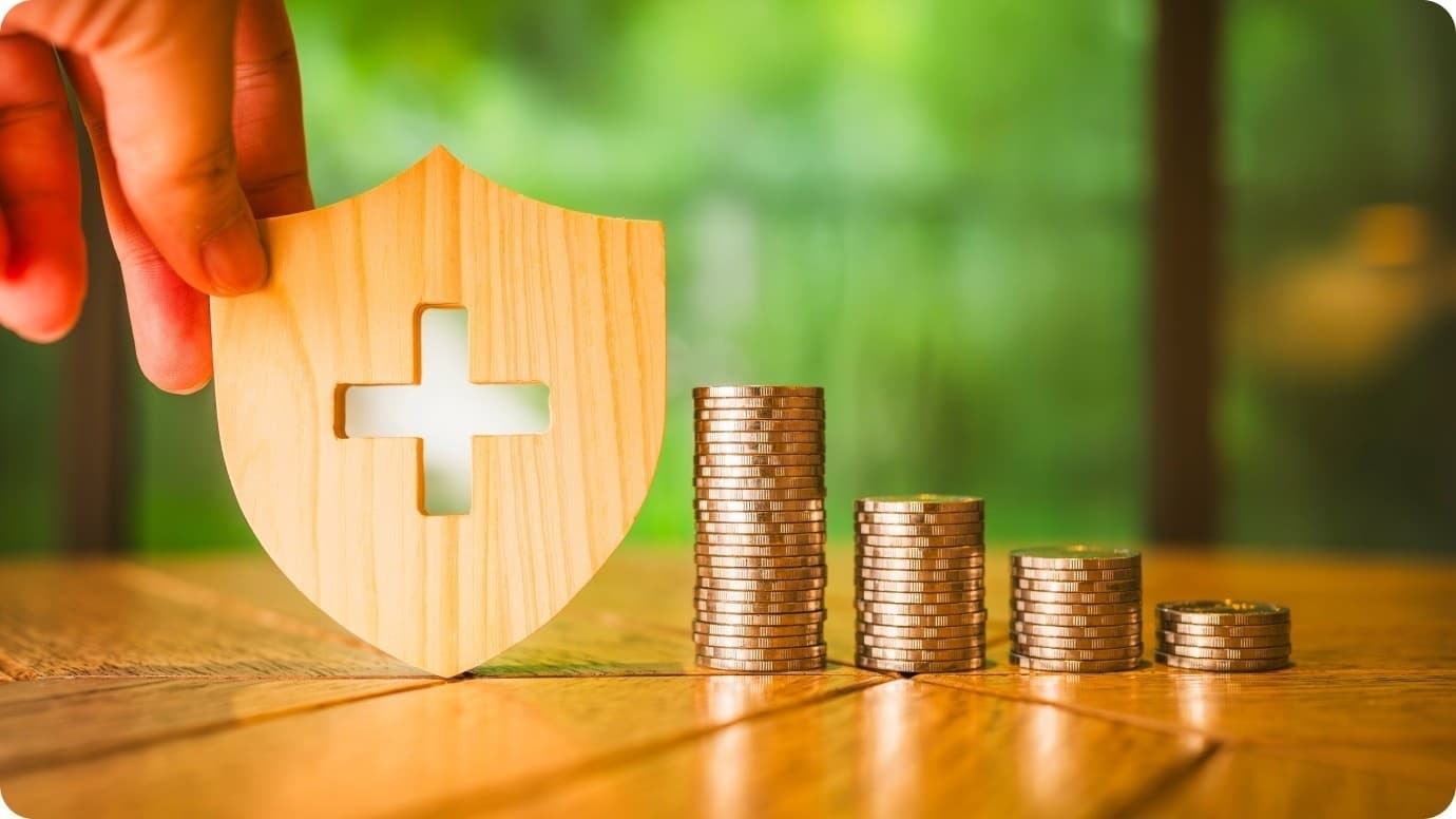 Hand holding a wooden shield with a medical cross beside stacked coins, symbolizing health funding.
